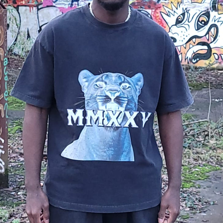 A man wearing a faded black graphic t-shirt featuring a blue jaguar print with the Roman numerals 'MMXXV' across the chest, set against a graffiti-covered outdoor wall.